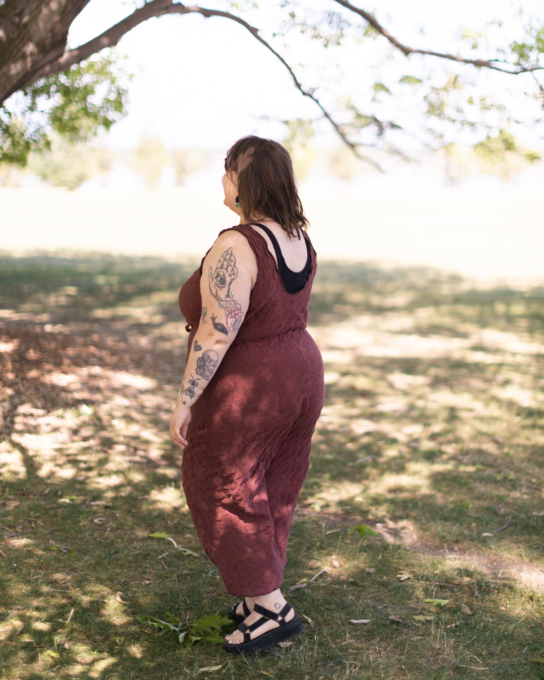 A photo of a woman wearing a True Bias Nova Jumpsuit in a mauve knit fabric standing in a shady spot outside under dappled light. 