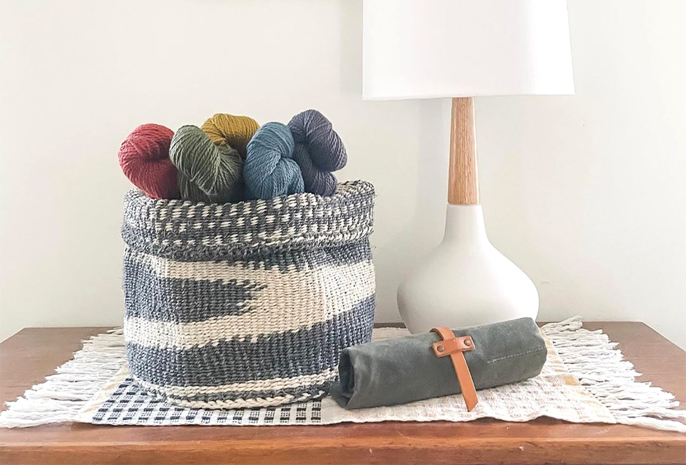 This is an image of a basket full of yarn sitting on top of a desk next to a lamp and a canvas roll for knitting needles