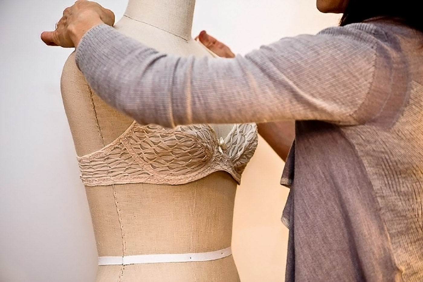 Close up of a woman's hands adjusting the straps of a white, lacy underwire bra on a dress form.