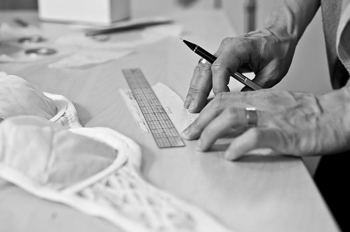 Close up of a woman's hands with a ruler and pencil, making adjustments to a sewing pattern piece for a bra. An unfinished, lacy bra lays on the table nearby.