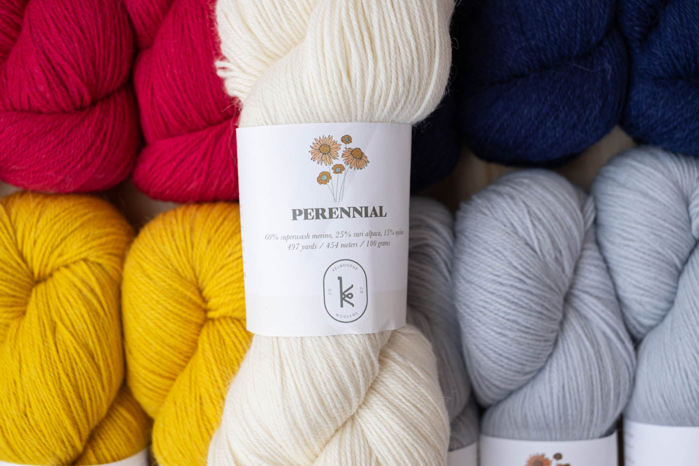 Kelbourne Woolens Perennial skeins in a lay flat photograph.  Color are hot pink, yellow, dark navy and light blue silver with on cream colored skein on the top of the pile.