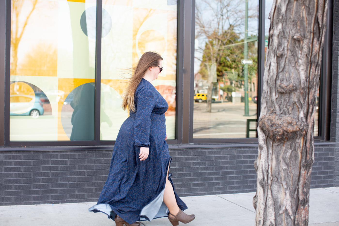 Shawna walking on a city sidewalk in front of a building.  Shawna is wearing a long navy flowy dress, that is blowing in the breeze.