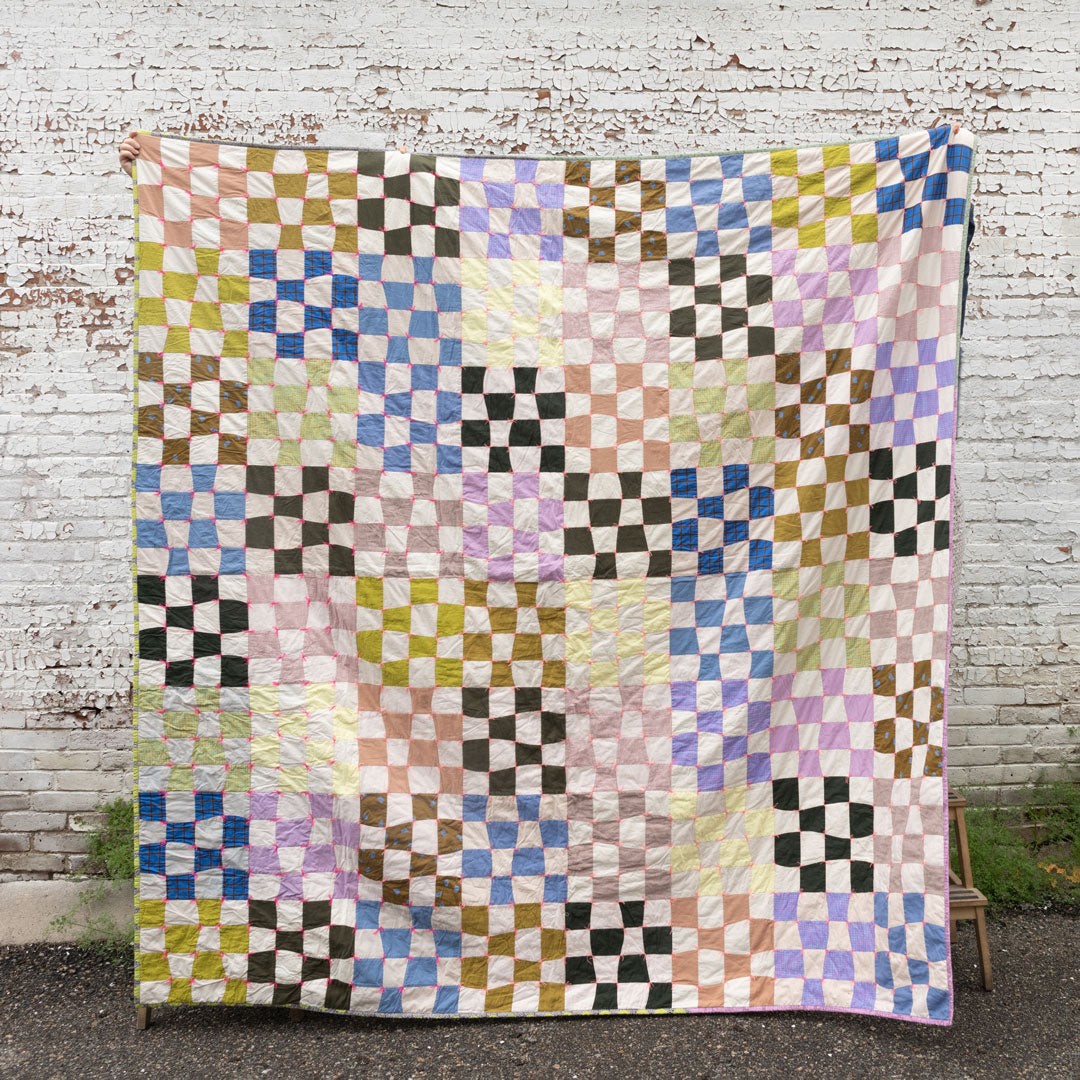 Shawna's Omega Quilt is held in front of a white brick wall. The quilt has a checkered design with an array of earthy colors