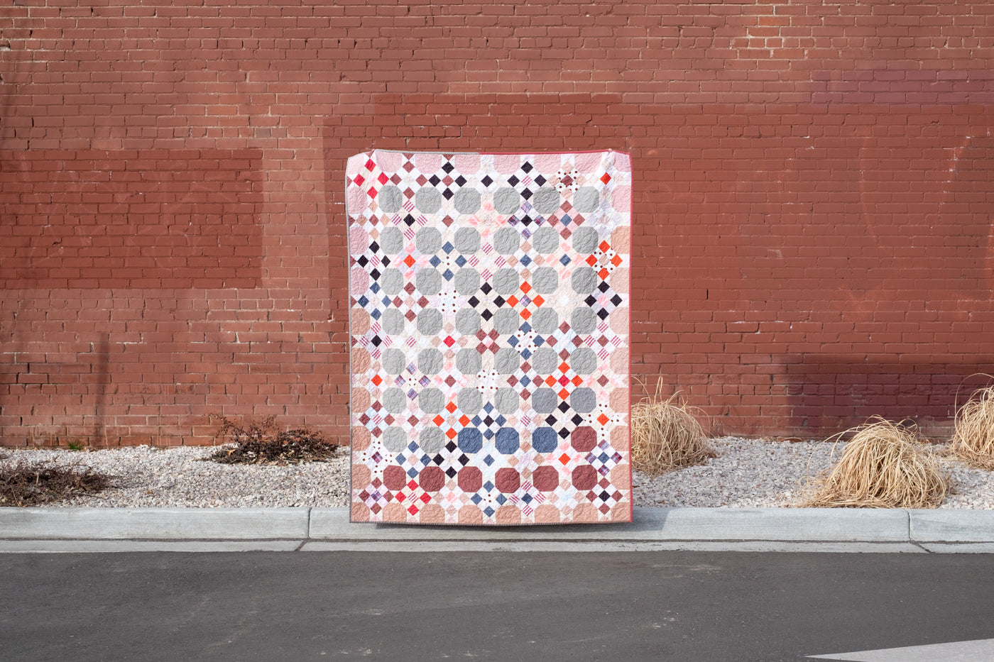 Photograph of scrap quilt, multi colored in front of a red brick wall.