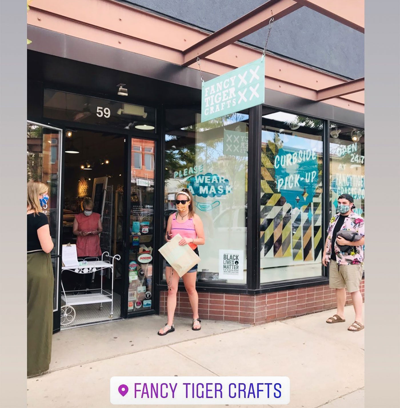 The storefront of Fancy Tiger Crafts. with a few customers outside standing 6 feet apart and wearing masks, waiting to pick up orders. An employees stands behind a makeshift counter at the door. A window mural says "Please wear a mask". 