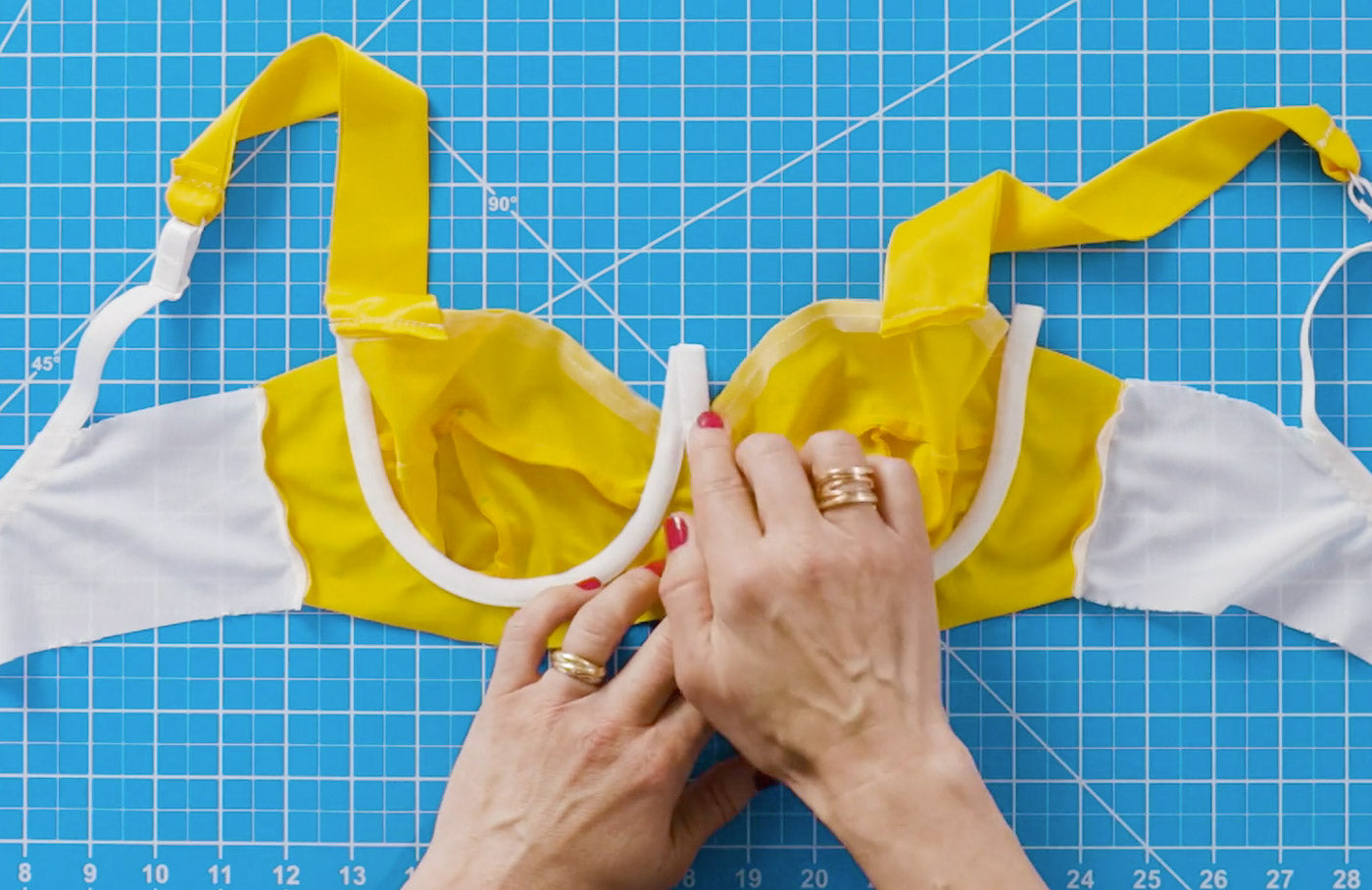 Hands placing the underwire casing onto an unfinished, handmade bra. The bra is color-blocked vibrant yellow and white and is laying on a bright aqua cutting mat. 