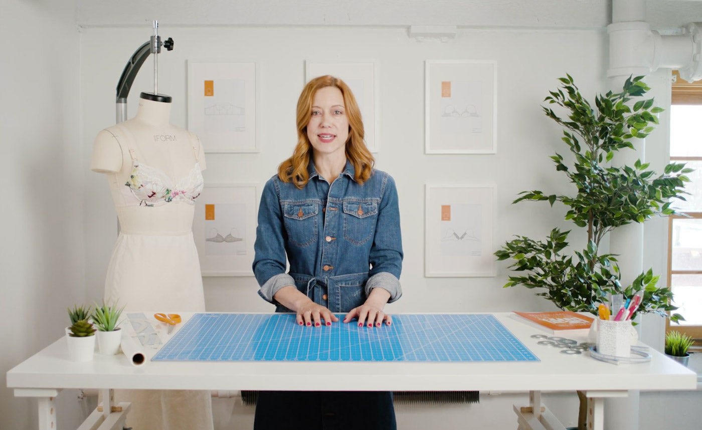 A still from Norma's bra-making video course, she stands behind a cutting table with some sewing tools, a dress form wears a floral bra next to her