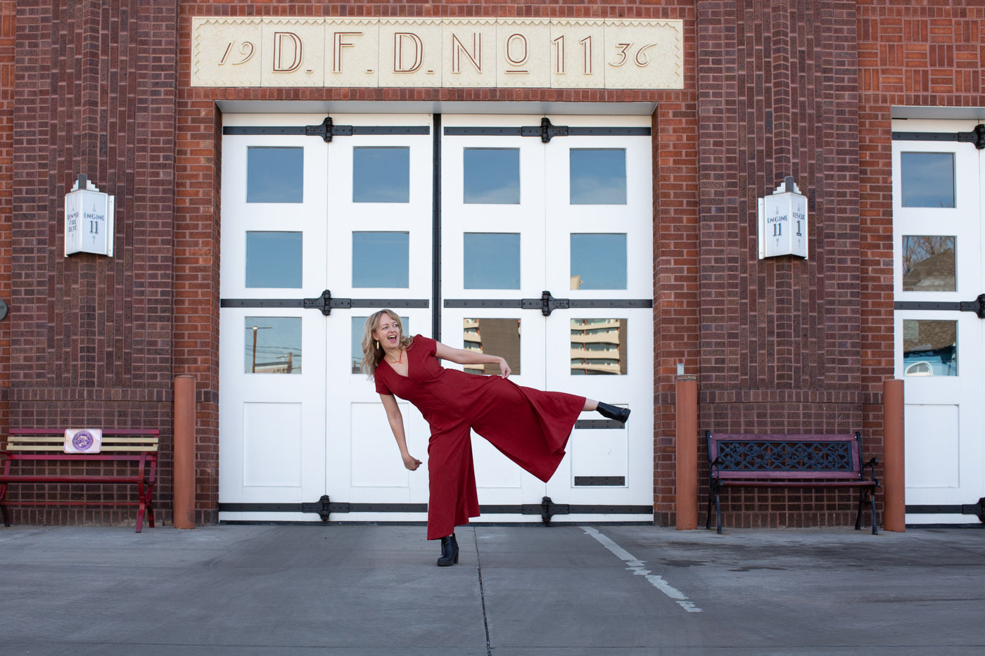 Amber wearing a red raw silk noil romper doing a high kick in front of a brick firehouse with white doors.