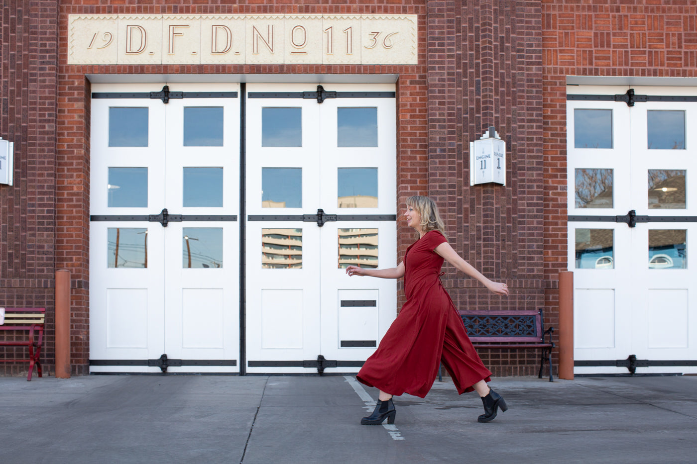 Amber taking large strides in her red silk noil romper in front of a red brick firehouse with large white doors.