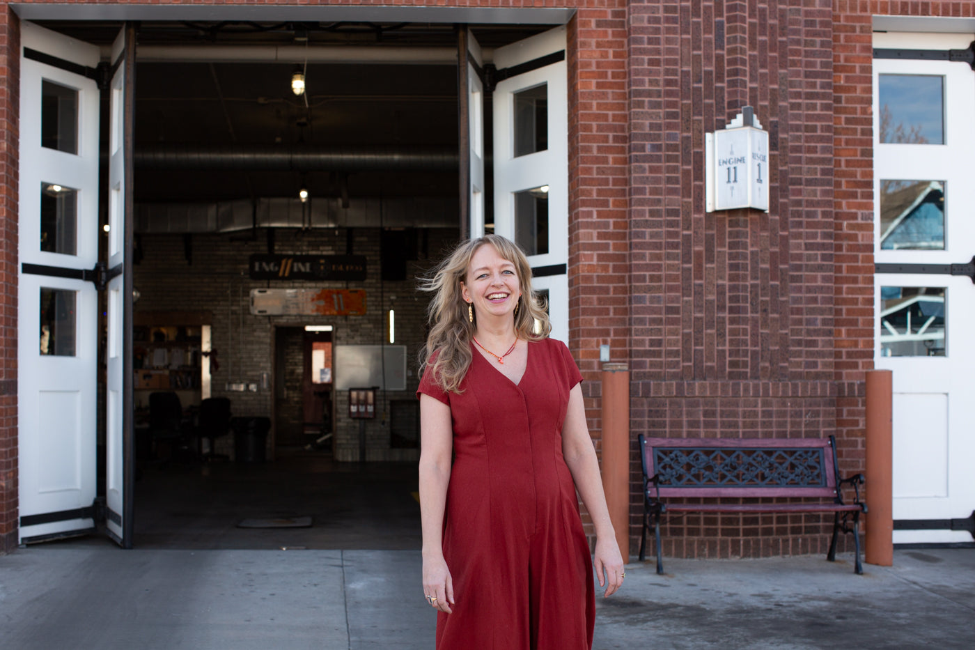 Amber in red silk noil romper in front of a red brick firehouse with white doors that are open into the firehouse.