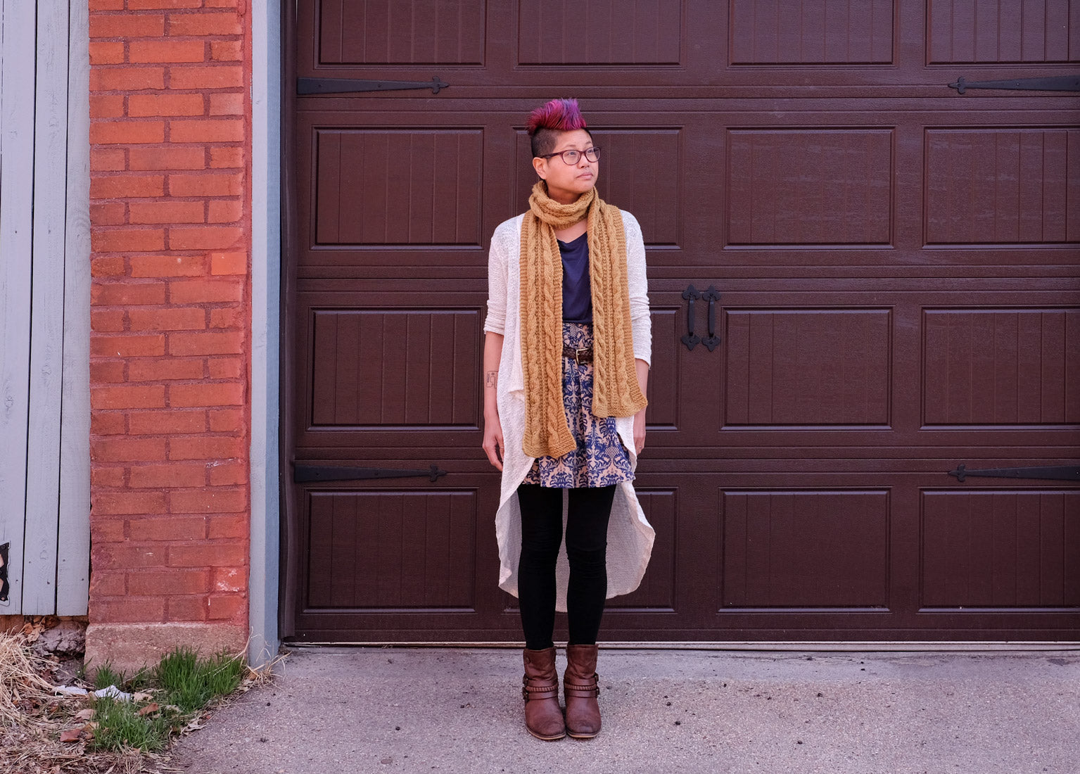 Woman with pink mohawk wearing mustard-yellow cabled scarf, standing in front of a dark brown garage door.