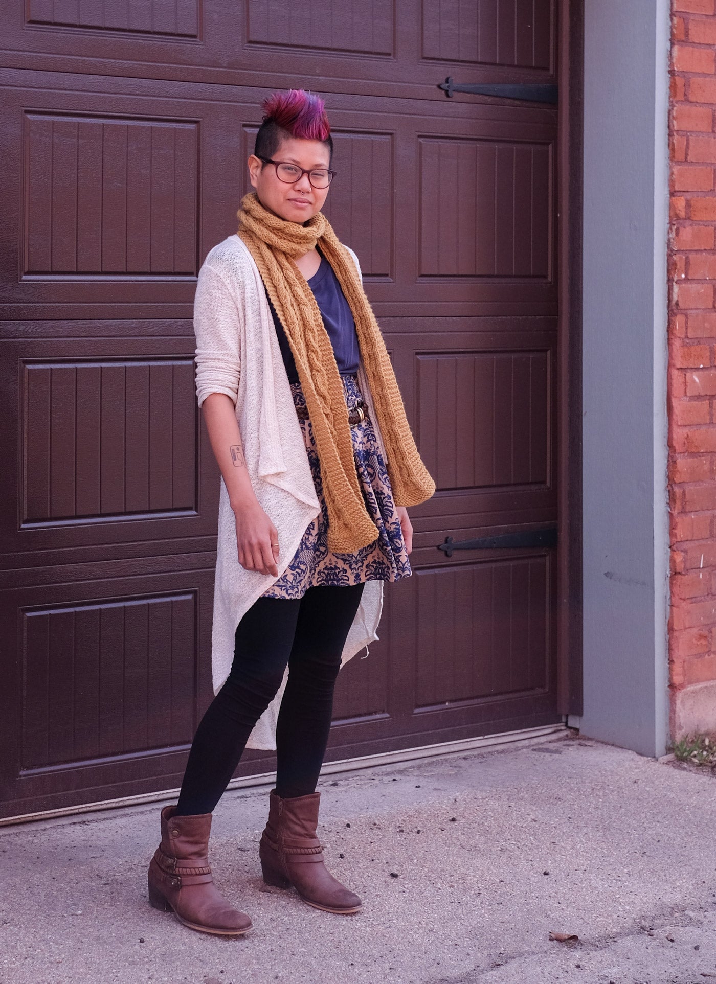 Woman with pink mohawk wearing mustard-yellow scarf, standing in front of a dark brown garage door.