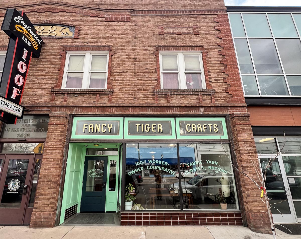 a view of the outside of the shop, Fancy Tiger Crafts Cooperative. The shop is in a historic brick building, and stands out in a joyful bright mint green. Gold leaf lettering in the upper windows shows the name of the shop, "FANCY TIGER CRAFTS", and the street level windows say "100% Worker-Owned Cooperative" and "Fabric, Yarn, Classes, Notions" in classic hand-painted lettering reminiscent of vintage shop fronts.