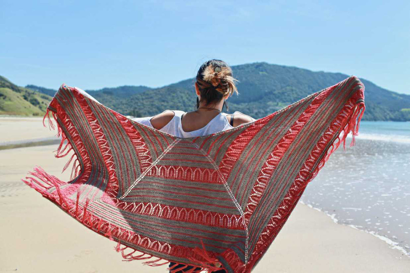 Outdoor photograph of a girl holding a brown/pink knitted shawl open.  Background is blue sky, mountain, ocean and beach.