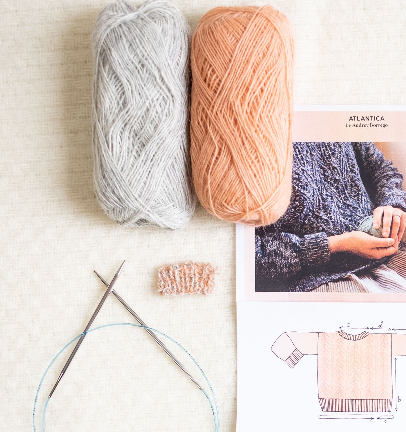 A ball of Light Ash Heather and a ball of Peach Einband yarn lay atop a printed page of the Atlantica Pullover pattern. A pair of stainless steel circular knitting needles and a small swatch of the two hues marled together lay next to the pattern page.  
