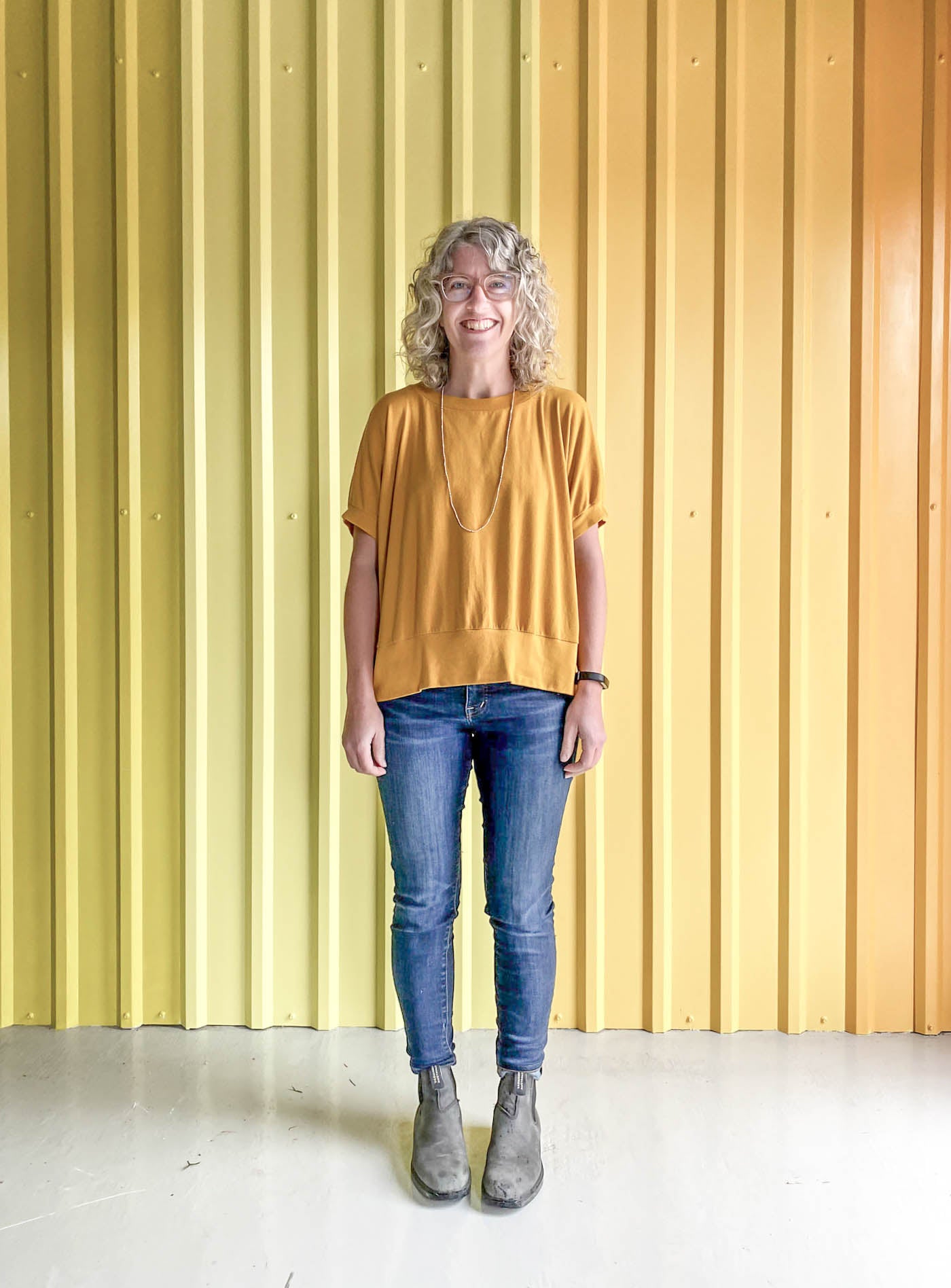 Jaime wearing a golden yellow Hosta tee, jeans and black boots, standing in front of a corrugated yellow ombre wall.