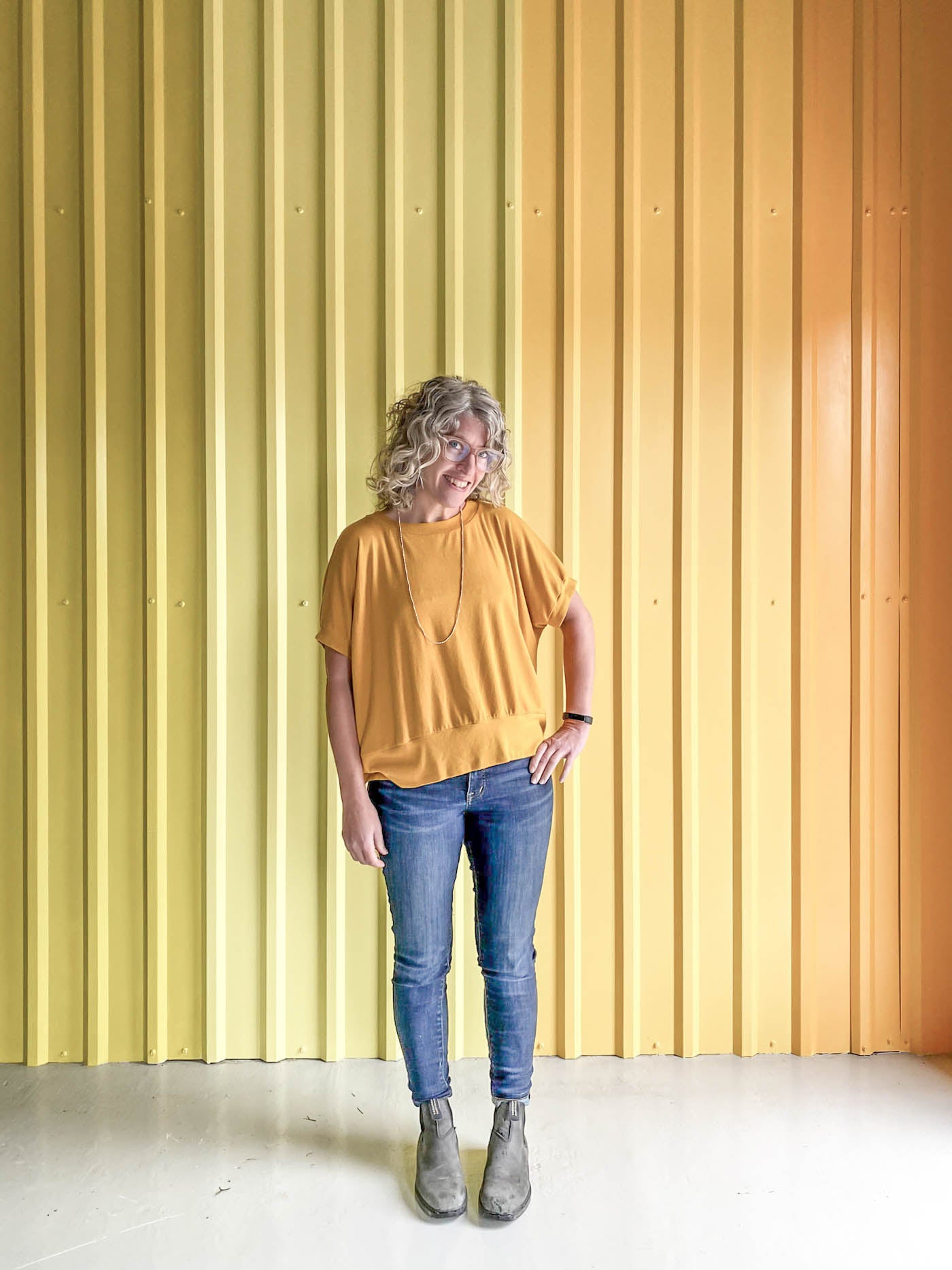 Jaime wearing a golden yellow Hosta tee, jeans and black boots, standing in front of a corrugated yellow ombre wall.