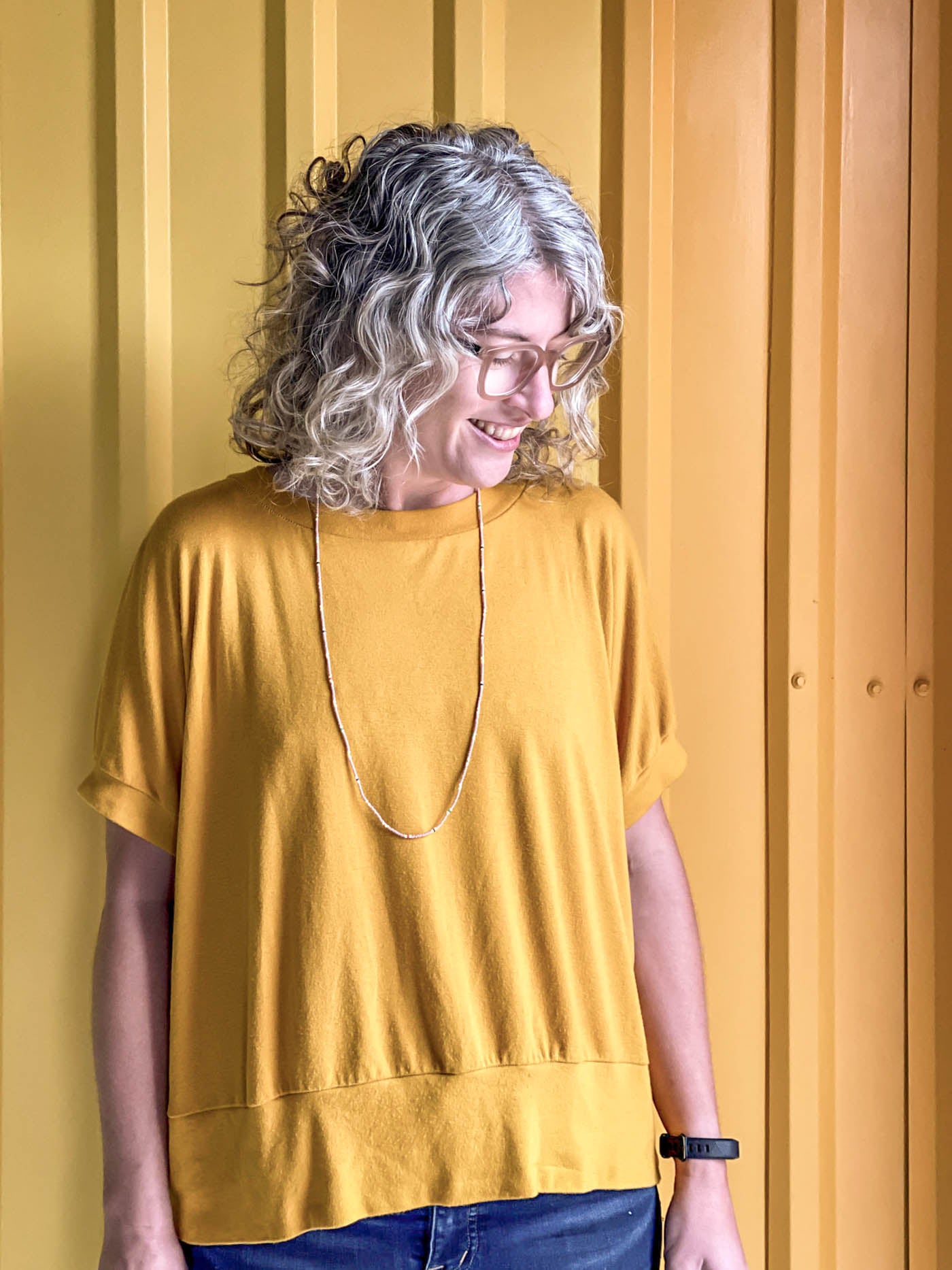 Close up of Jaime looking down, wearing a golden yellow Hosta tee, standing in front of a corrugated yellow ombre wall.