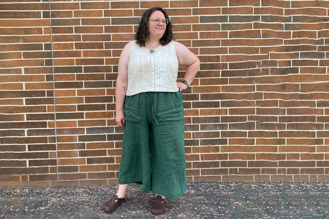 Kim wearing her pair of Soline Culottes in Nomad Linen Twill in the color Juniper . She wears a white stripe tank top and is standing in front of a brick wall