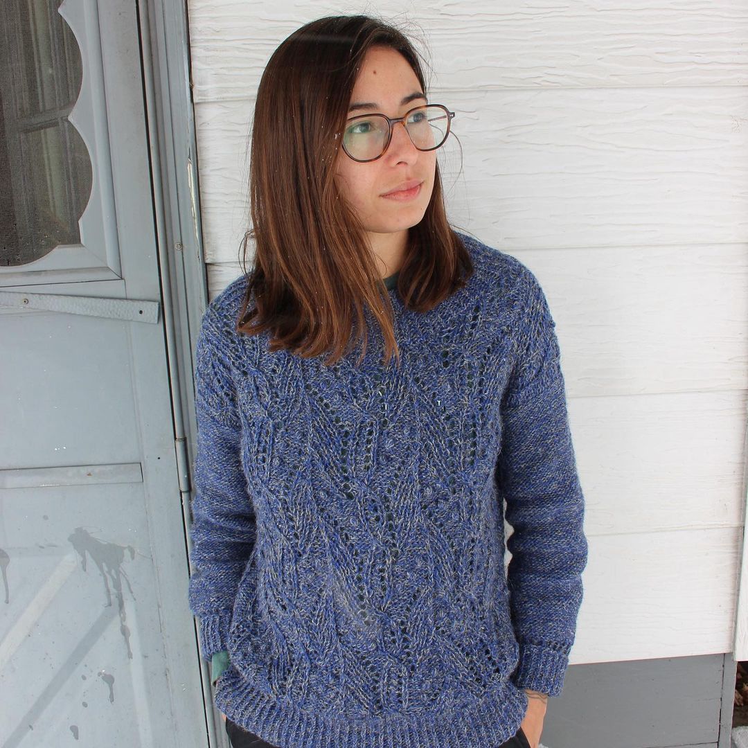 Luna stands outside near a porch door, wearing the denimy blue Atlantica pullover she recently knit. 