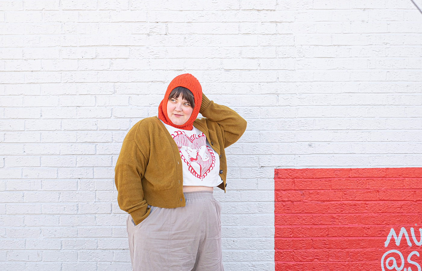 Marta wearing a red balaclava in front of a grey wall with an accent square that is also red