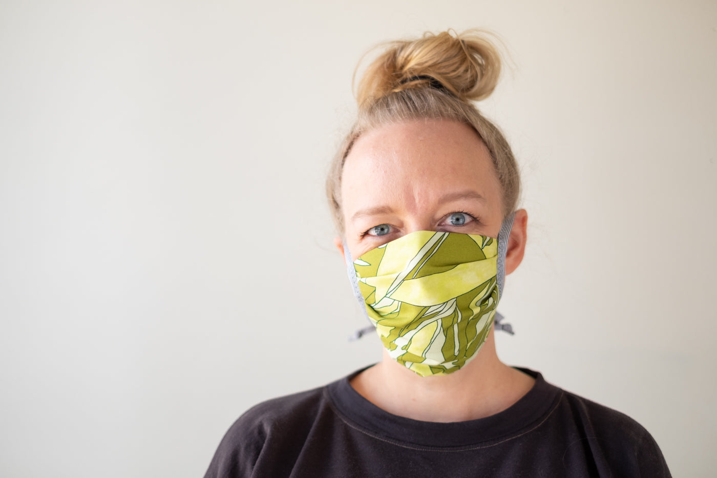 A closely cropped photo of Amber against a white background, she is wearing a fabric mask in an abstract green print and her hair is in a top-knot.