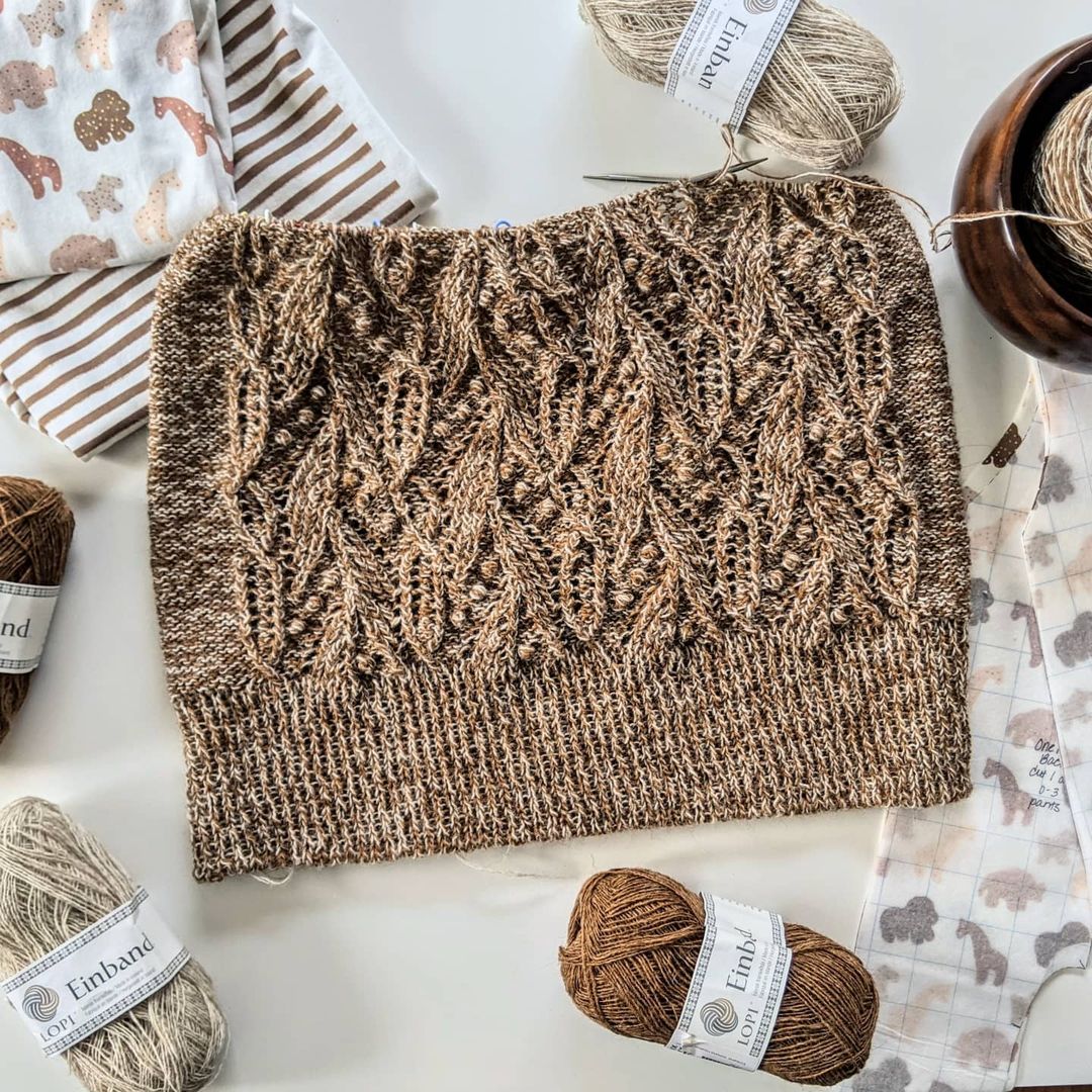 An in-progress Atlantica sweater in warm almond hues lays flat on a white surface. It is surrounded by it's yarn balls, and a cute sewing project in similar colors that is also in-progress, using adorable fabric featuring an animal cookie print.