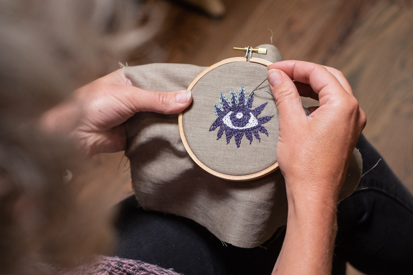 This is an image of a persons hands holding an embroidery hoop with an eye stitched on the fabric. 