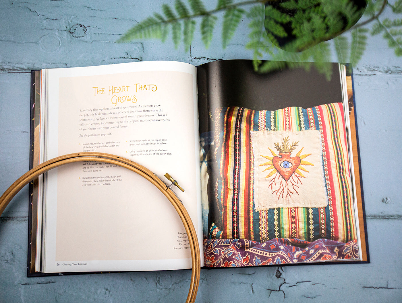 This is an image of a book opened up to a page with heart stitched embroidery project along with a wooden embroidery hoop and a plant. 