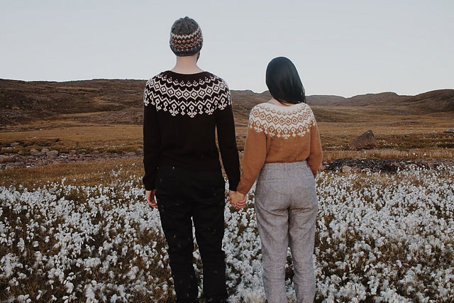 Photo Credit to Ravelry boy and girl standing in a field with their backs facing camera.  Couple is holding hand showing their knitted sweaters.  The man is wearing a knitted hat with a black and white sweater and black pants.  The women has her dark brown hair to the side showing the back of her mustard yellow and white knitted sweater wearing medium grey tweed pants.