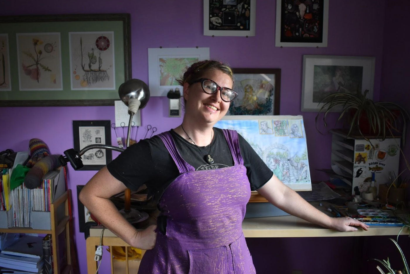 Pink stands in her office. There is a variety of framed pictures hanging on a purple wall behind her. Pink is wearing a purple sundress and a black t-shirt under the dress. 