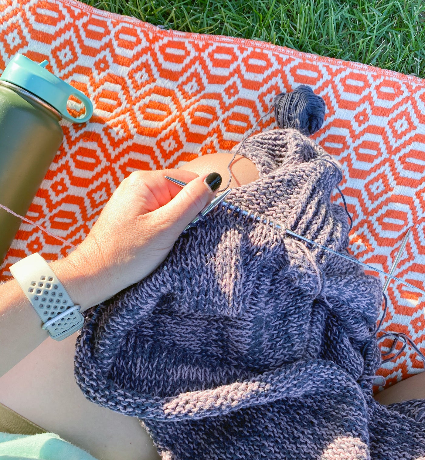 A close-up of someone's hands knitting a cotton garment, they sit crosslegged on a poppy-red and white patterned picnic blanket on lush green grass, with a green metal water bottle laying next to them. 
