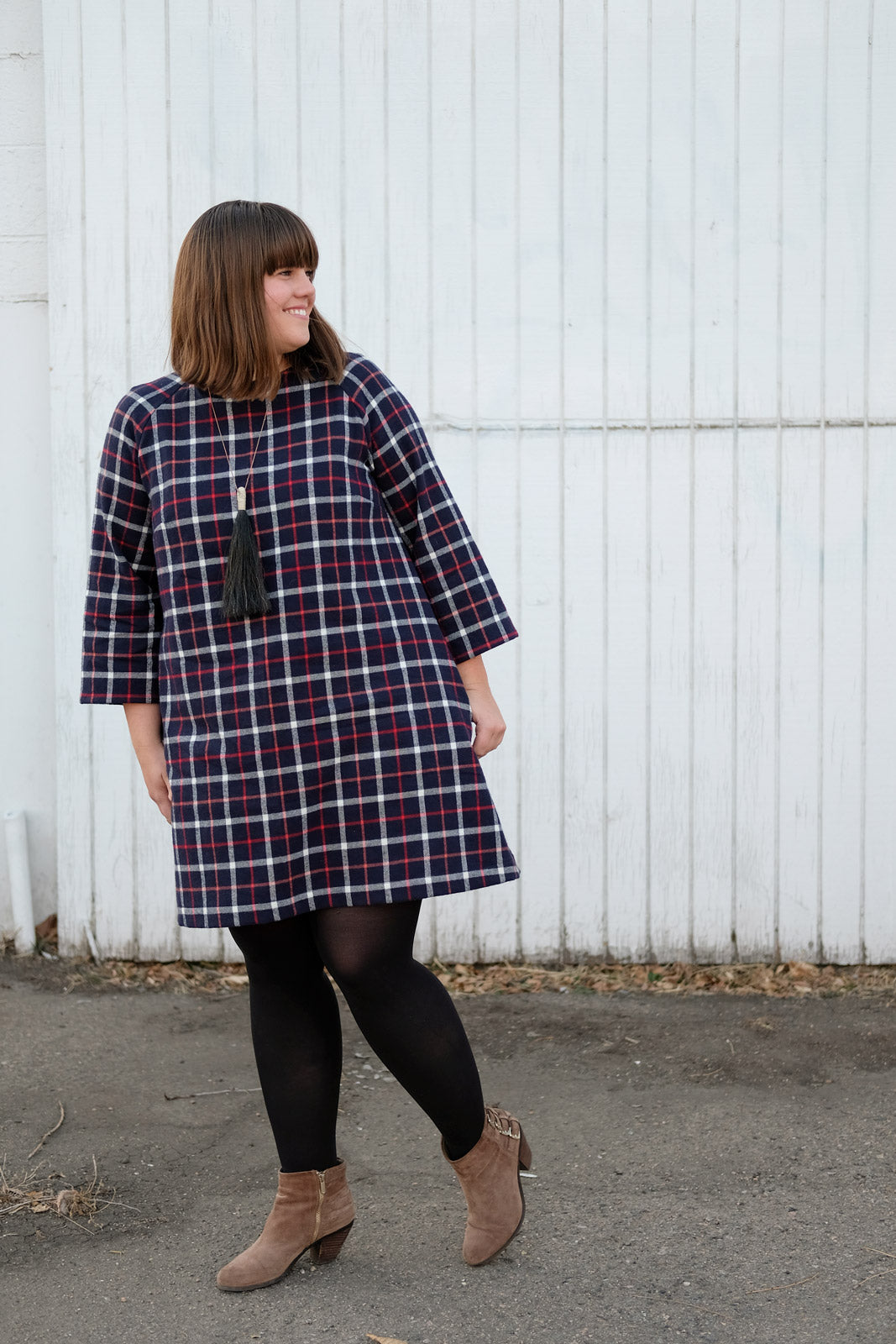 Kaylee in her blue plaid Avid Seamstress Raglan Dress in front of a white wall looking off to the side