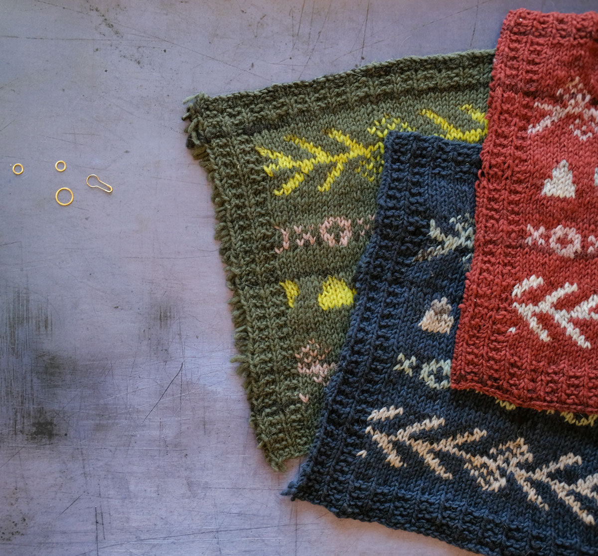 Three colorwork knitting swatches, red, navy and green, stacked on a grey background.