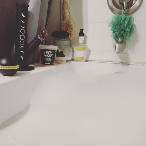 A point of view shot from someone relaxing in a bathtub with bubbles. There is a black Ooze Saturn Globe Grinder and black bong on the side of the tub. There are also some toiletry bottles and a green loofah hanging from the faucet.