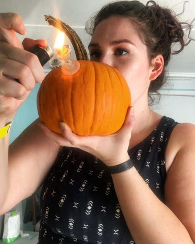A young white, brunette female is lighting the bowl of her pumpkin bong and taking an inhale. It is a small pumpkin with an Ooze flower bowl inserted on the opposite side of her face.