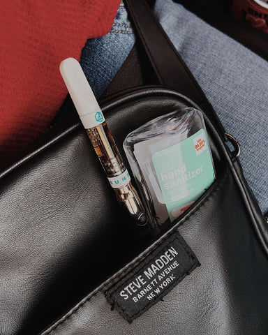 A close-up shot of a woman's black leather crossbody bag. There is an Ooze Slim Twist Vape Pen and travel size bottle of hand sanitizer sticking out of the outer pocket.