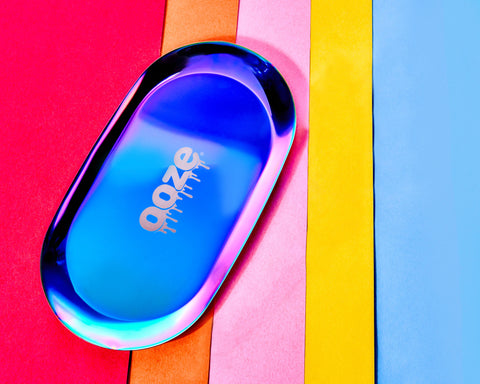 A rainbow Ooze Aura rolling tray is laying on an angle on top of bright, striped background.