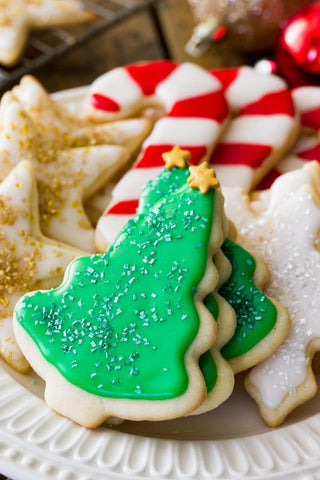 Photo credit to the Sugar Spun Run blog (https://sugarspunrun.com/easy-sugar-cookie-recipe/). A pile of Christmas sugar cookies are arranged on a plate. Front and center are green Christmas trees with gold stars on top, gold star cookies are off to the left, and candy cane cookies are in the back.