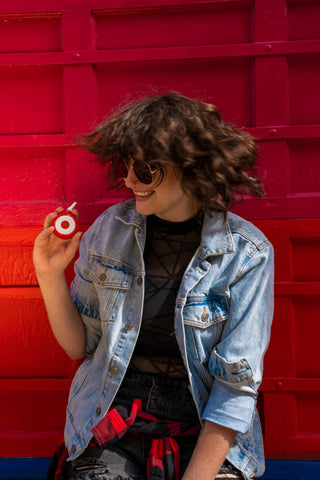 A young brunette white woman in a jean jacket and sunglasses is dancing with her red Ooze Movez Speaker Vape in her hand.