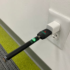 A black Ooze Slim Twist Vape Battery is charging. It is connected to the Ooze Smart USB Charger and in a white cube adapter in the wall. The battery light is glowing green and the charger light glows red to indicate it is charging.