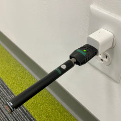 A black Ooze Slim Twist Vape Battery has just completed its charge cycle. It is plugged into the Ooze Smart USB Charger and a white cube adapter, plugged into the wall. The battery light is turned off and the charger light glows green to indicate charge is complete.