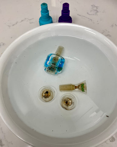 Four Ooze bowls are soaking in a white bowl filled with clear liquid to remove the debris. A blue Glyco bowl, regular Ooze flower bowl, and two Armor Bowl inserts are there, with the Armor Bowl silicone sitting on the counter.