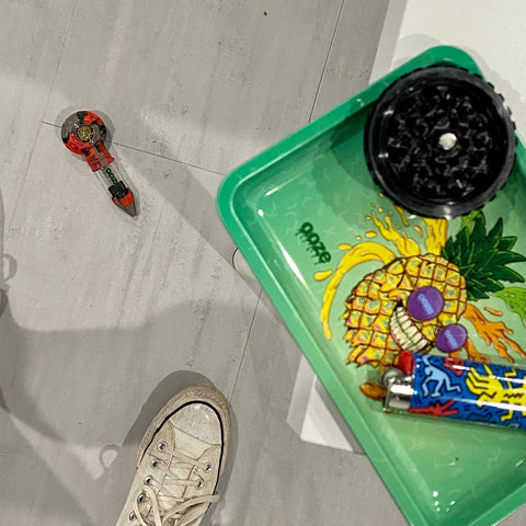 A shot looking down at the floor. The Ooze after midnight Bowser Travel Pipe is on the floor facing up with a fully packed bowl. A white converse sneaker is at the bottom of the frame. On the edge of the white table is a small metal Mr. Pineapple Ooze rolling tray, a black Saturn grinder bottom piece, and a blue K. Haring lighter.