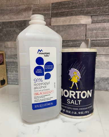 A bottle of 91% isopropyl alcohol is on a white granite countertop next to a large cannister of Morton table salt. The backsplash is a rugged gray tile in a mix of different shades.