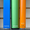 A blue Ooze Quad battery, green Novex battery, and orange Slim Twist vape pen battery are all lined up together on a black shelf. The shot is a close up of the authentication code at the base of each battery.