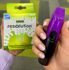 A girl with a pink manicure holds her galaxy purple Ooze Booster vape next to a 100ct box of Micro Swabs