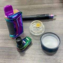 A rainbow Ooze Duplex 2 vaporizer is sitting on a gray table with the base detached. An onyx atomizer is attached with an open jar of concentrate and an Ooze Hot Knife next to it