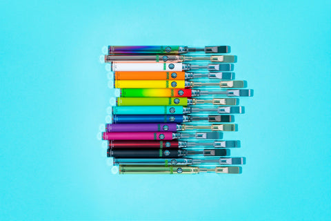 All of the original Ooze Slim Twist Pen 1.0 vape batteries are laying flat on an aqua background arranged in rainbow order.