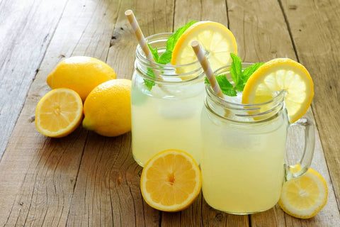 Two glass mason jar mugs filled with THC-infused lemonade with lemon slices and garnish on the rim, surrounded by lemons